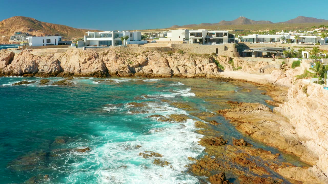 Elevated frame at Chileno Beach showing white buildings along the cliff, a small cove of turquoise water, foamy waves over exposed rocks, and no visible people.