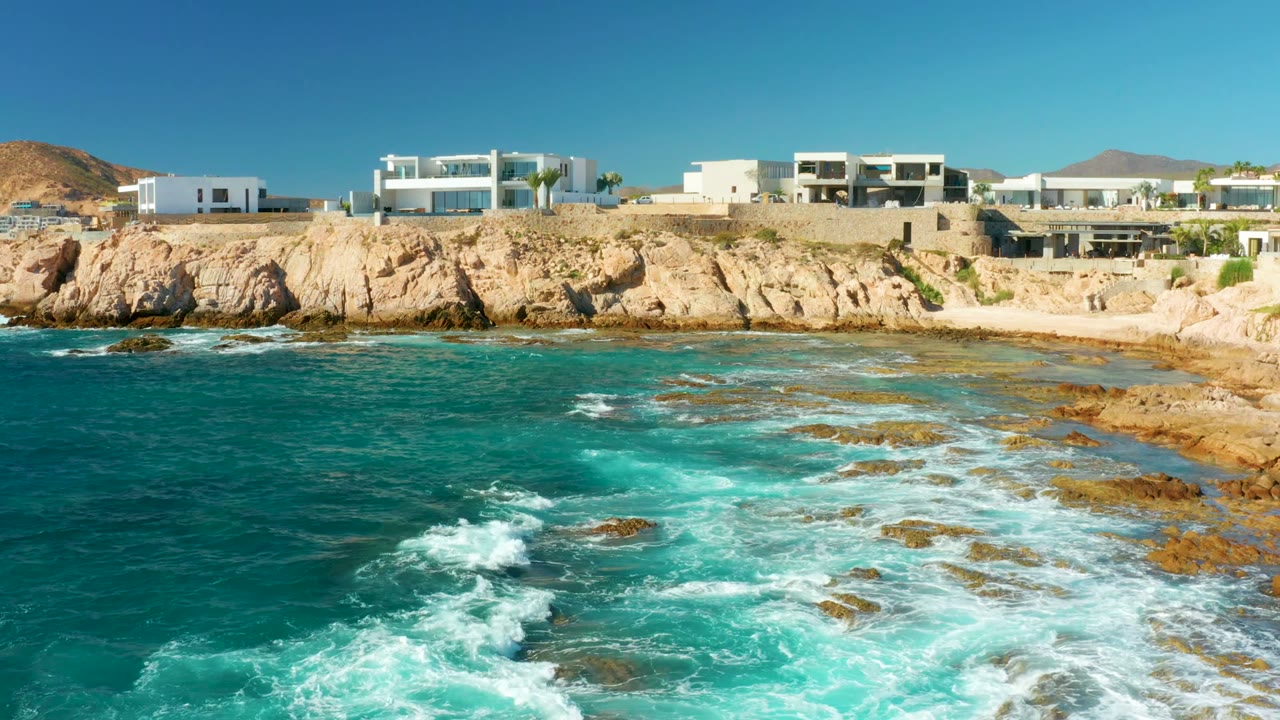 Opening frame at Chileno Beach, Los Cabos shows turquoise water and white foam over rocks below pale cliffs with white modern villas and no visible people.