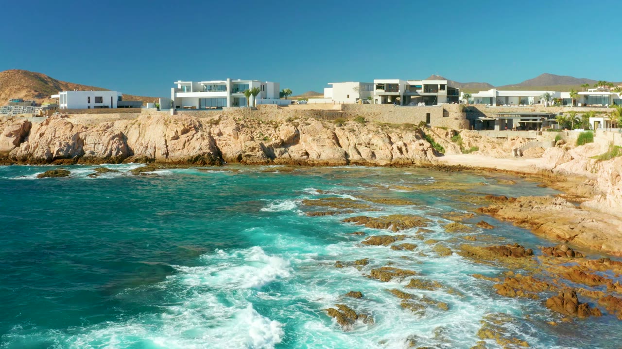 At 0:02, the wave foam gathers nearer the center of the rocky coast, with modern white villas above the Chileno Beach cliff and no visible people.