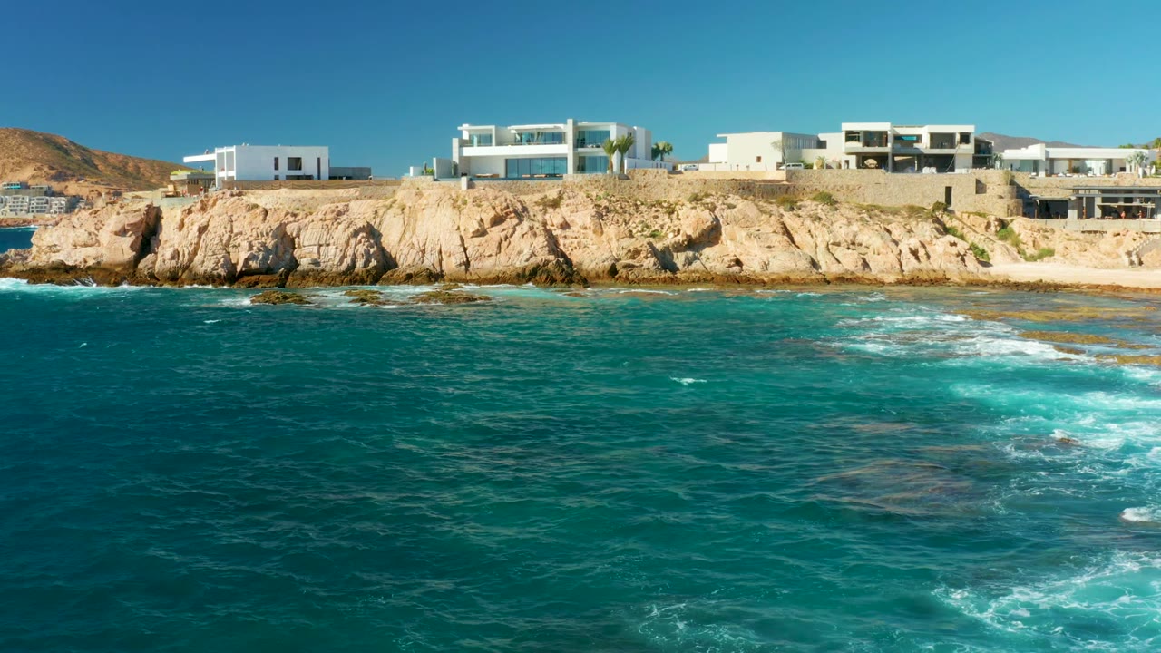 Opening frame of Chileno Beach in Los Cabos with turquoise water in the foreground, tan rocky cliff across the middle, white buildings above, and no visible people.