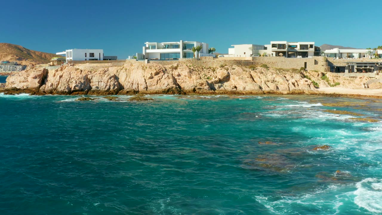 Chileno Beach surf increases at 0:02 as white foam spreads across rocks in the foreground, with tan cliffs and white buildings behind, and no visible people.