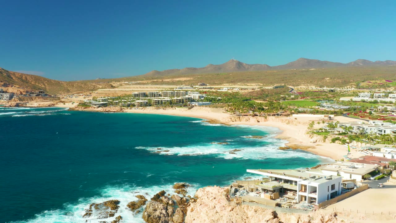 At 0:00, an elevated view over Chileno Beach in Los Cabos shows turquoise water breaking over rocks, a sandy cove, beachfront buildings, brown mountains, clear sky, and no visible people.