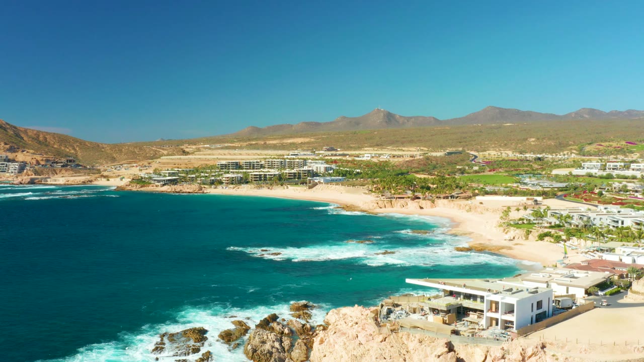 Wide aerial coastline with waves rolling toward sand and buildings below mountains.