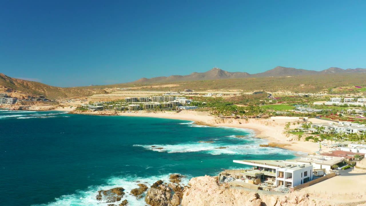 At 0:02, foamy waves pull across the dark turquoise water near Chileno Beach rocks while pale sand, modern beachfront buildings, distant mountains, clear sky, and no visible people stay in view.