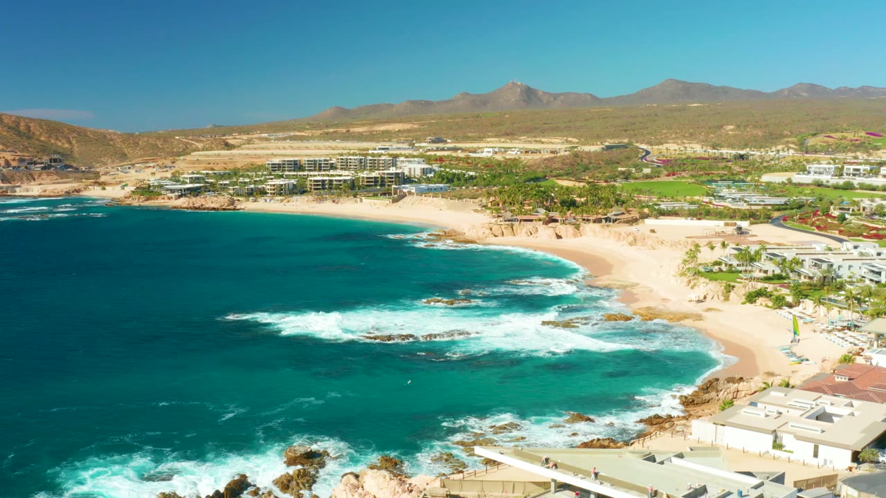 Opening frame over Chileno Beach in Los Cabos, with turquoise water on the left, waves breaking near sand and rocks, coastal buildings at lower right, mountains in the distance, and no visible people.