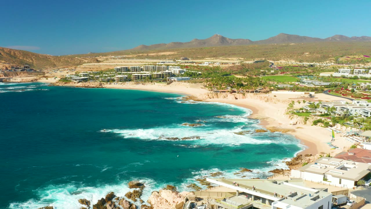 Waves break across rocks beside a sandy beach with buildings and mountains beyond.