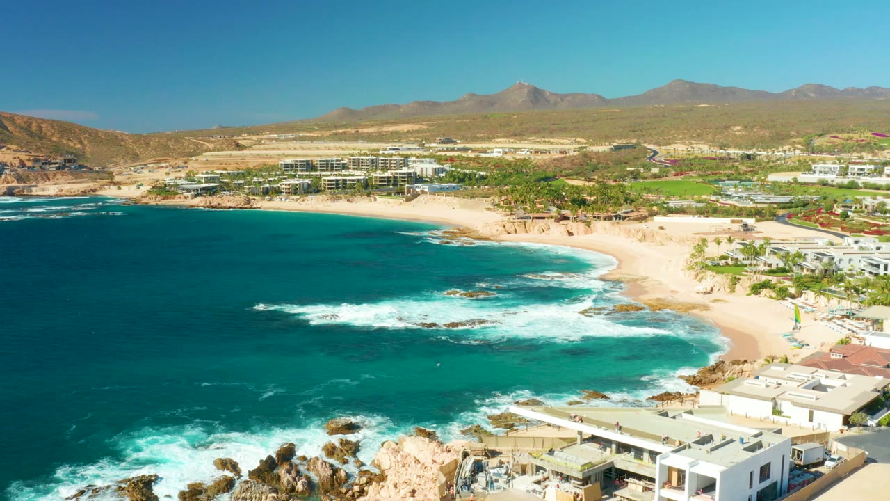 At 0:02, the aerial view holds the Chileno Beach shoreline with deep blue water at left, foamy surf near the beach, pale buildings in the lower right, and no visible people.