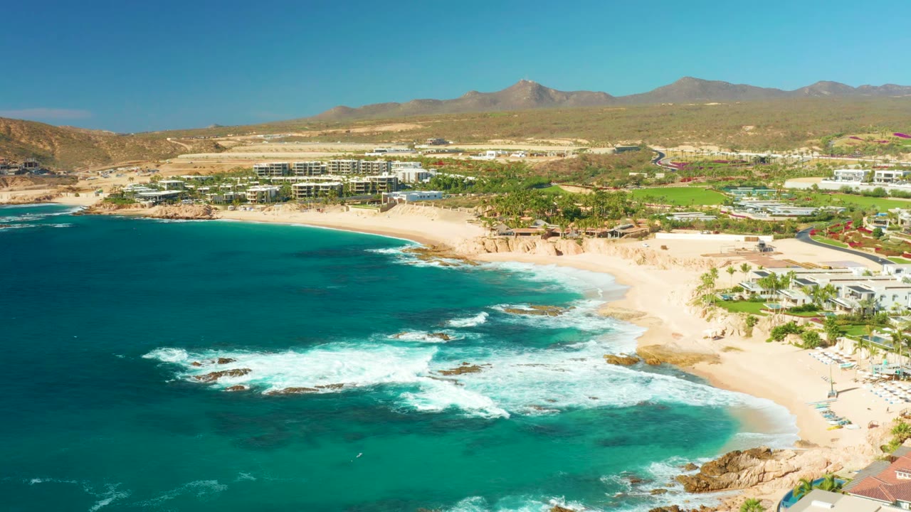Aerial frame over Chileno Beach in Los Cabos showing turquoise water, foamy waves on offshore rocks, sandy shoreline, coastal buildings, dry mountains, and no visible people.