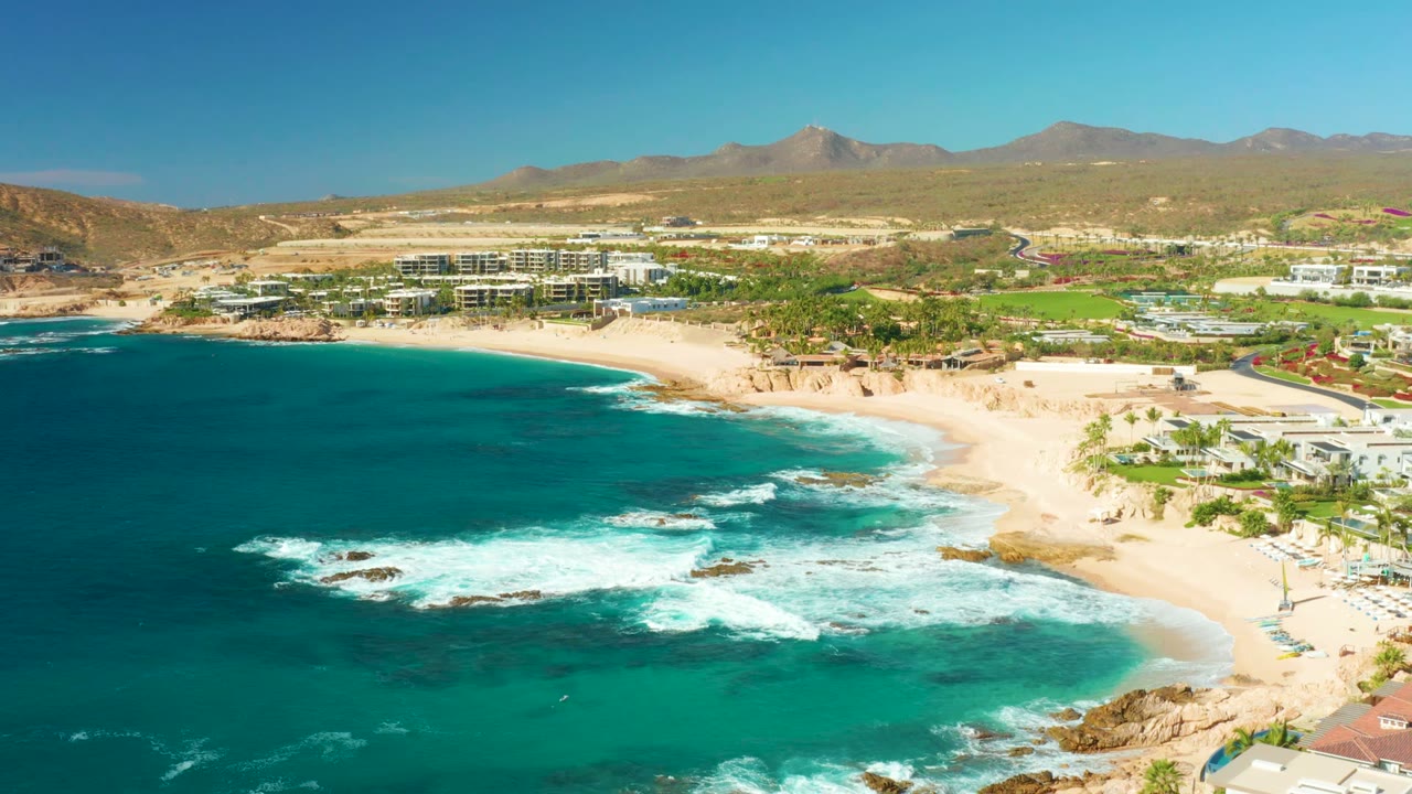 Wide aerial coastline with waves breaking across rocks beside a sandy beach and coastal development.