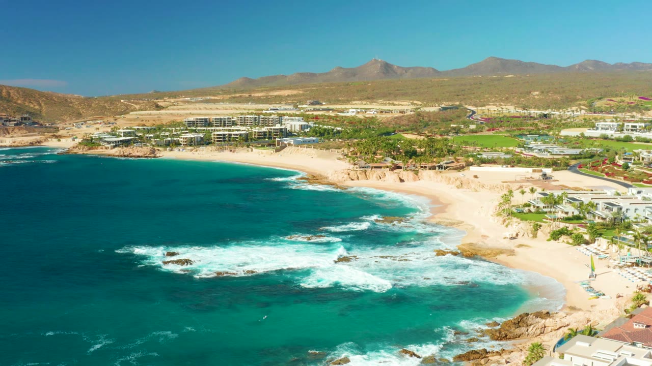 Wide aerial frame of Chileno Beach showing white waves advancing toward the sand, rocky patches in turquoise water, low coastal buildings, desert hills, distant mountains, and no visible people.