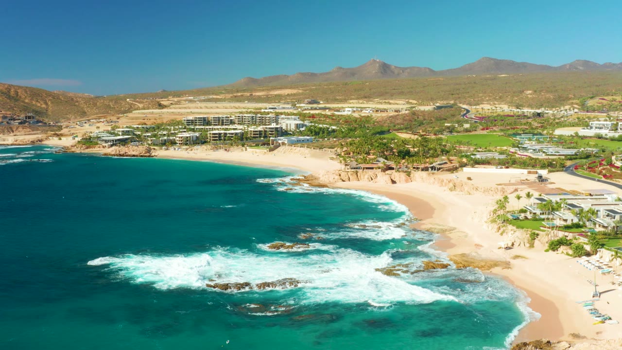 Aerial frame over Chileno Beach in Los Cabos showing turquoise water on the left, white surf over rocks, a sandy shore with small beachgoers, and dry mountains beyond coastal buildings.
