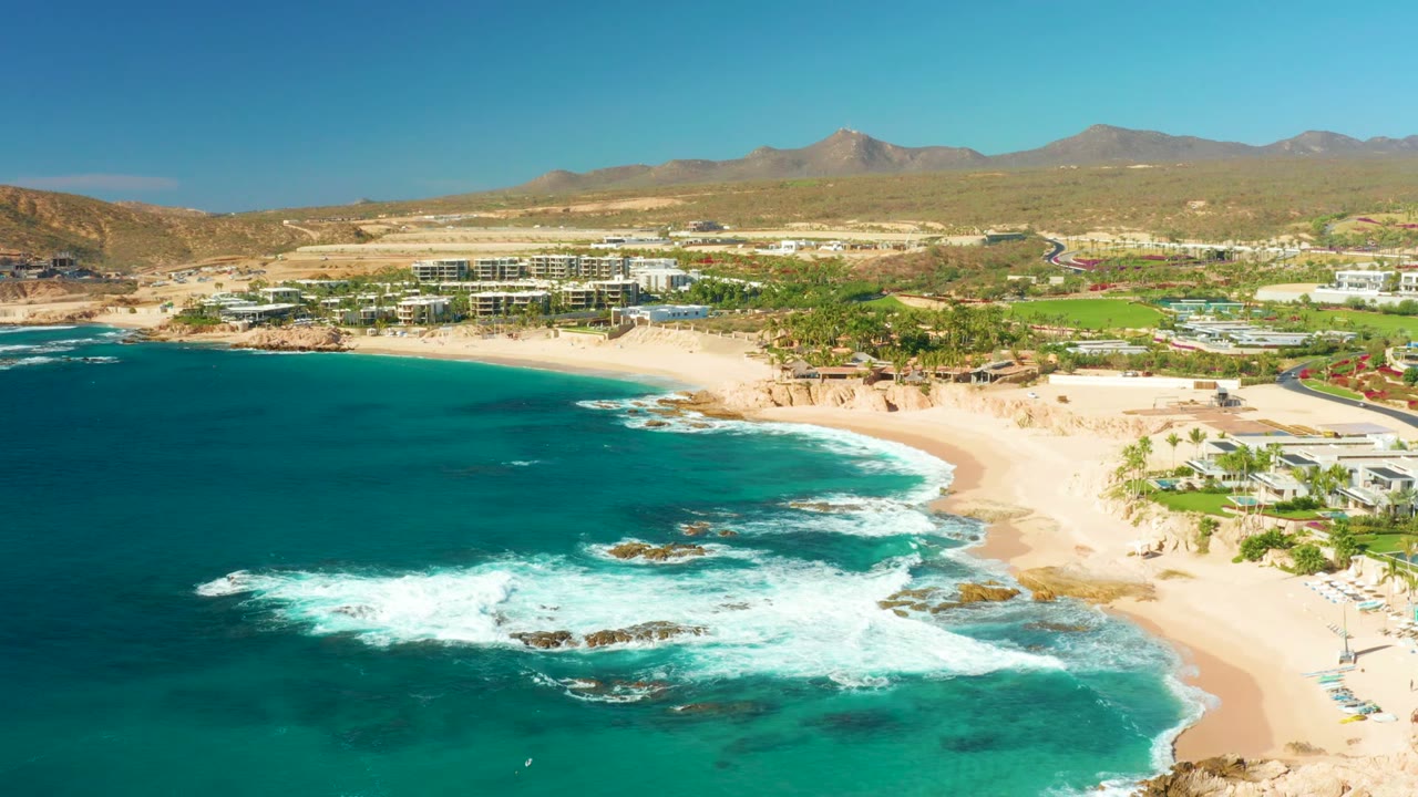 Wide aerial beach view with waves breaking around offshore rocks.