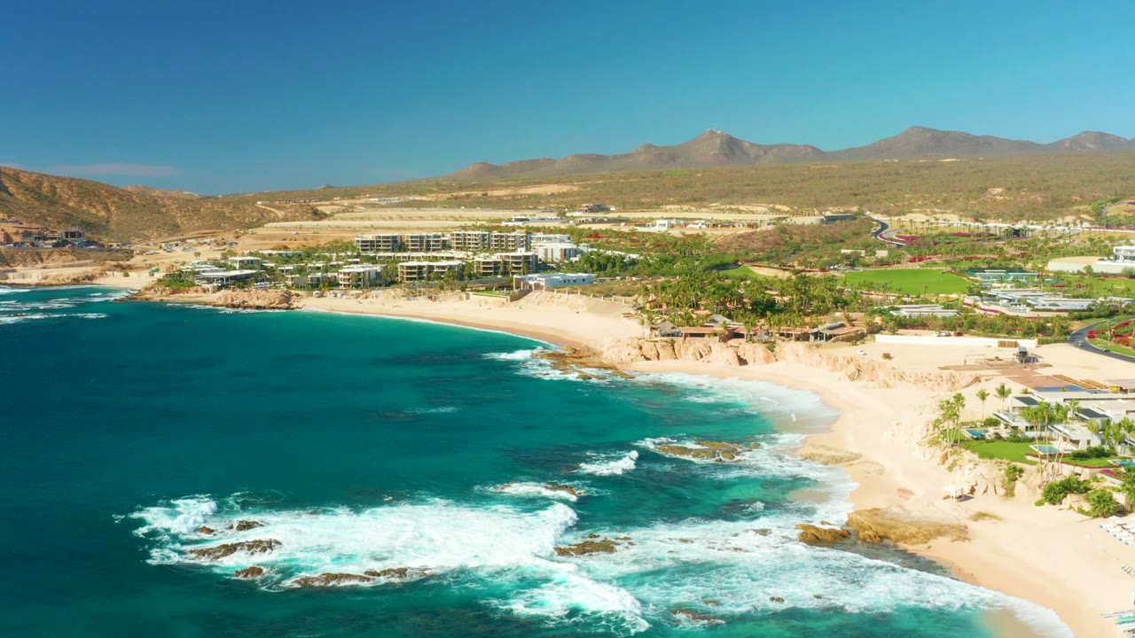Opening aerial frame over Chileno Beach in Los Cabos, with turquoise water and white surf in the foreground, a sandy curve at right, resort buildings inland, mountains beyond, and no visible people.