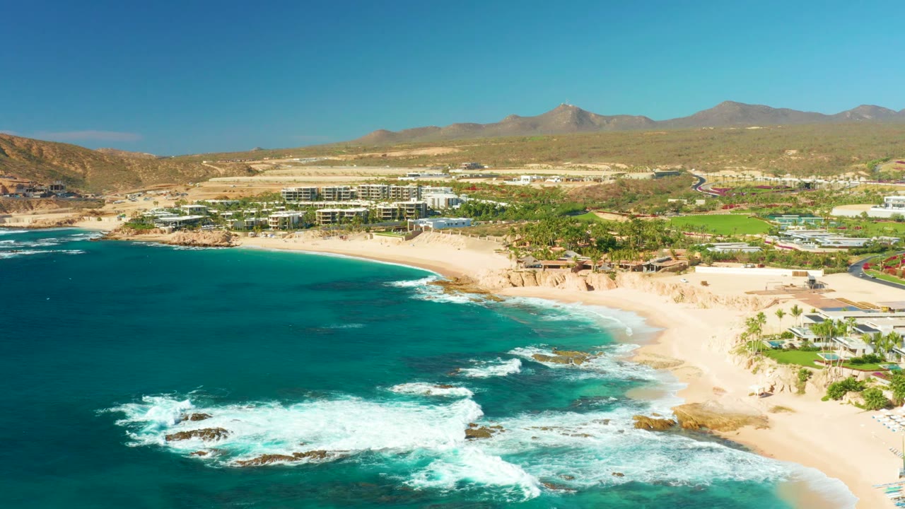 White waves spread over rocks near a curved beach with resort buildings inland.