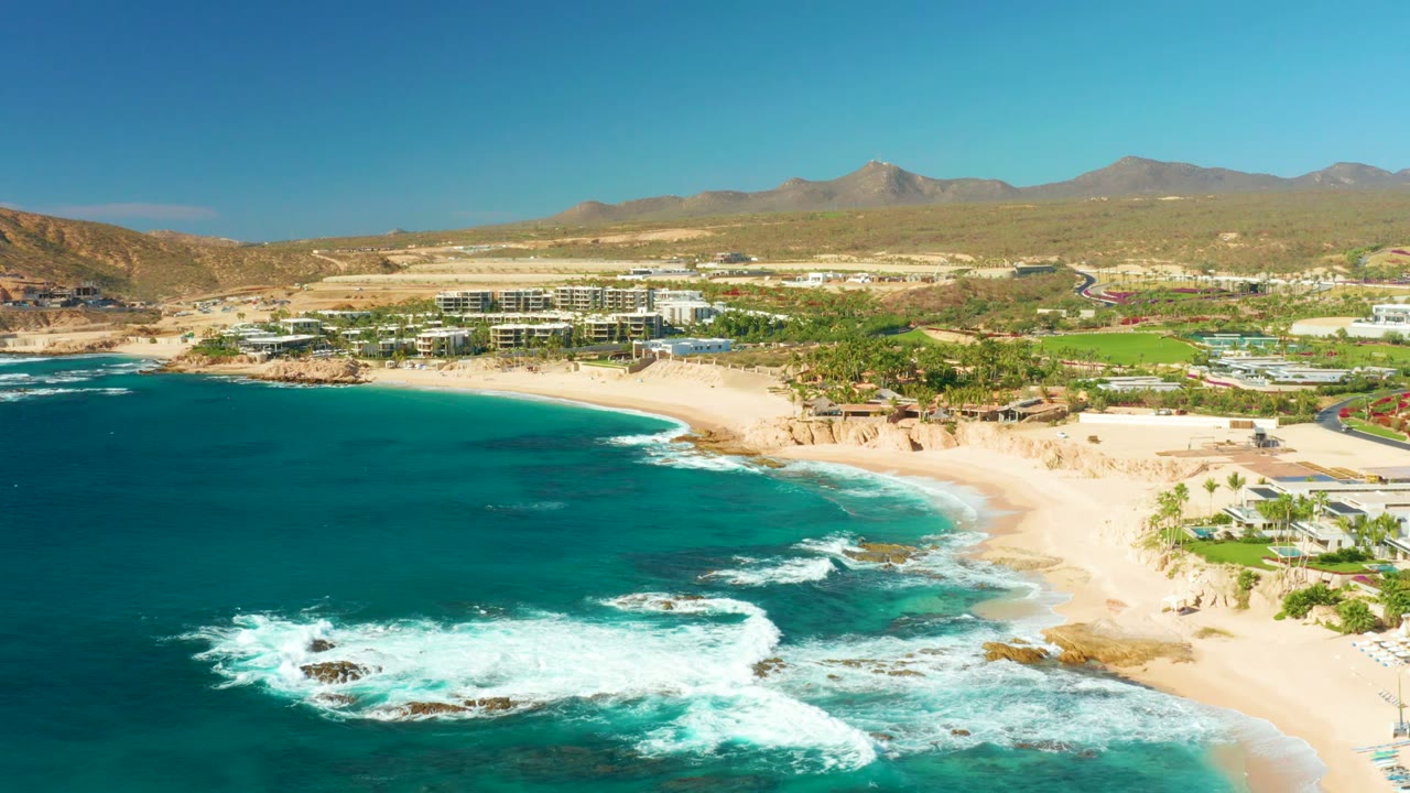 At 0:02, the Chileno Beach aerial holds a broad coastline view with surf breaking lower in frame, the sandy beach sweeping right, coastal buildings and green landscaping inland, distant mountains, and no visible people.