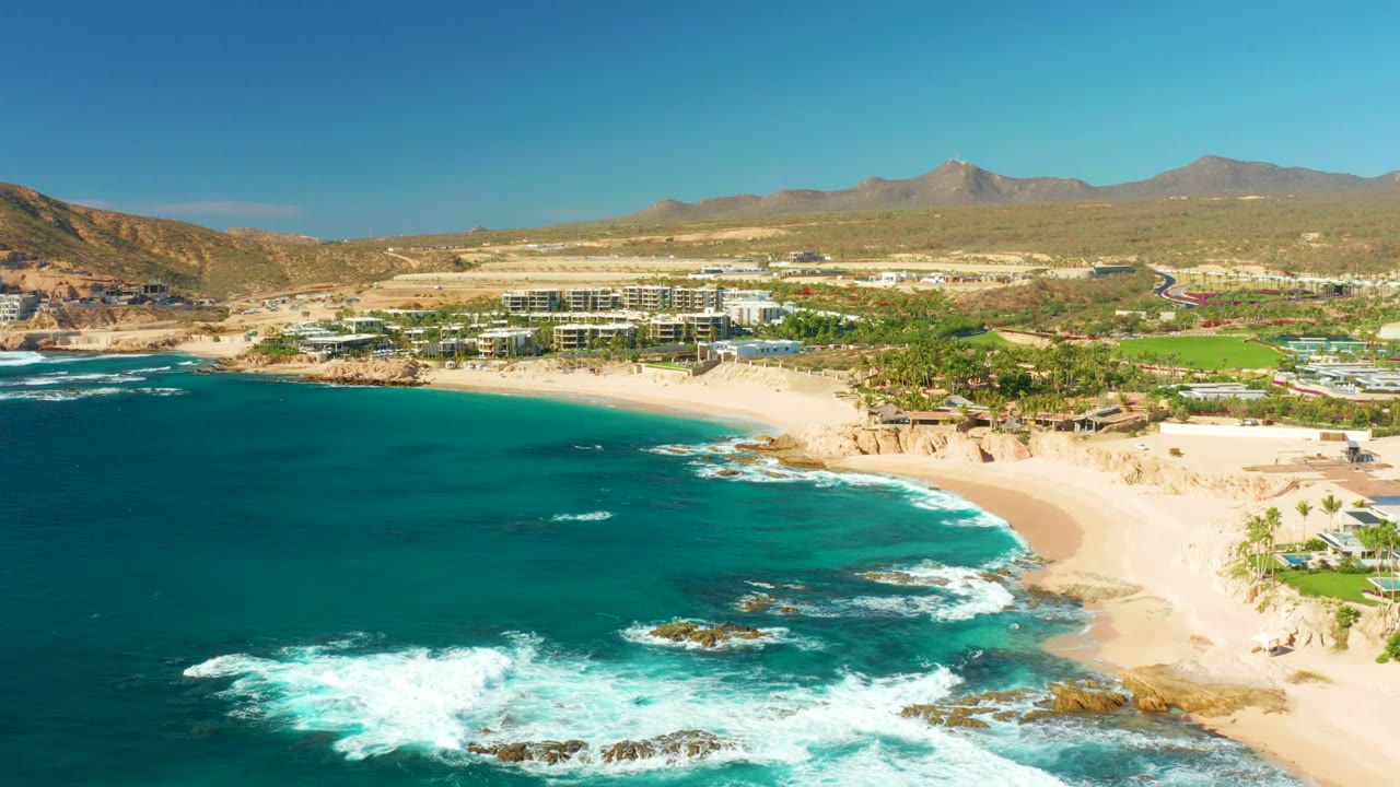 Aerial frame over Chileno Beach in Los Cabos showing turquoise water breaking over rocks beside a wide sandy beach, resort buildings inland, desert hills on the horizon, and no visible people.