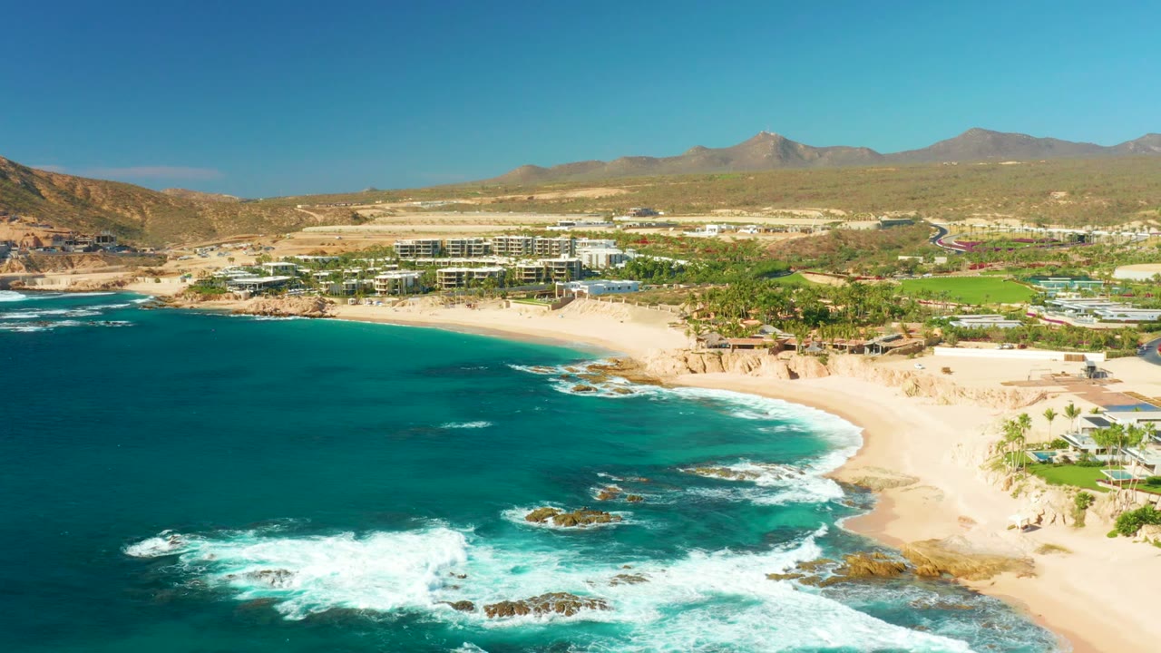 Aerial Chileno Beach frame showing blue ocean water, a band of white surf along rocks, the sandy shoreline curving toward resort buildings, and no visible people.