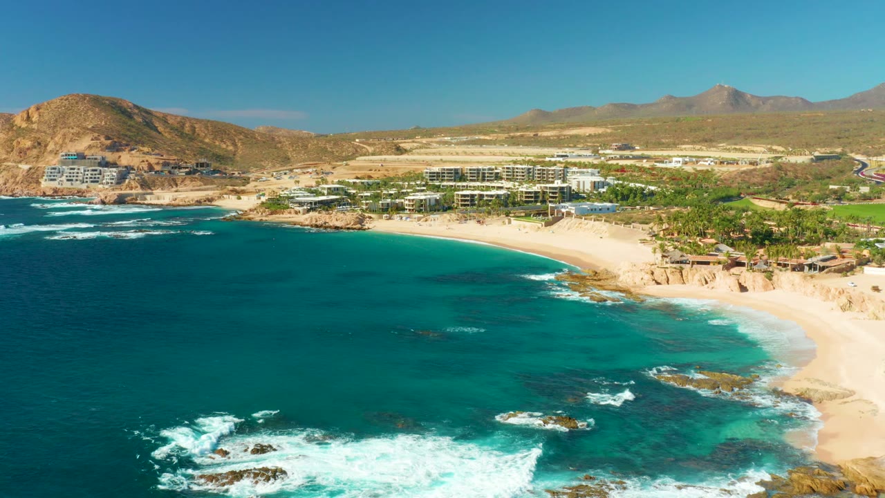 Aerial view over Chileno Beach in Los Cabos, with turquoise water and white surf in front of sand, coastal buildings, dry hills, and no visible people.