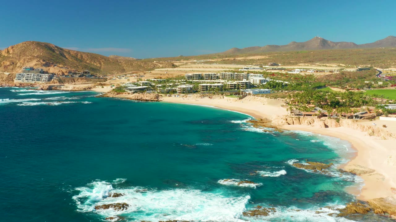 White waves break over rocks below a curved sandy beach and coastal buildings.
