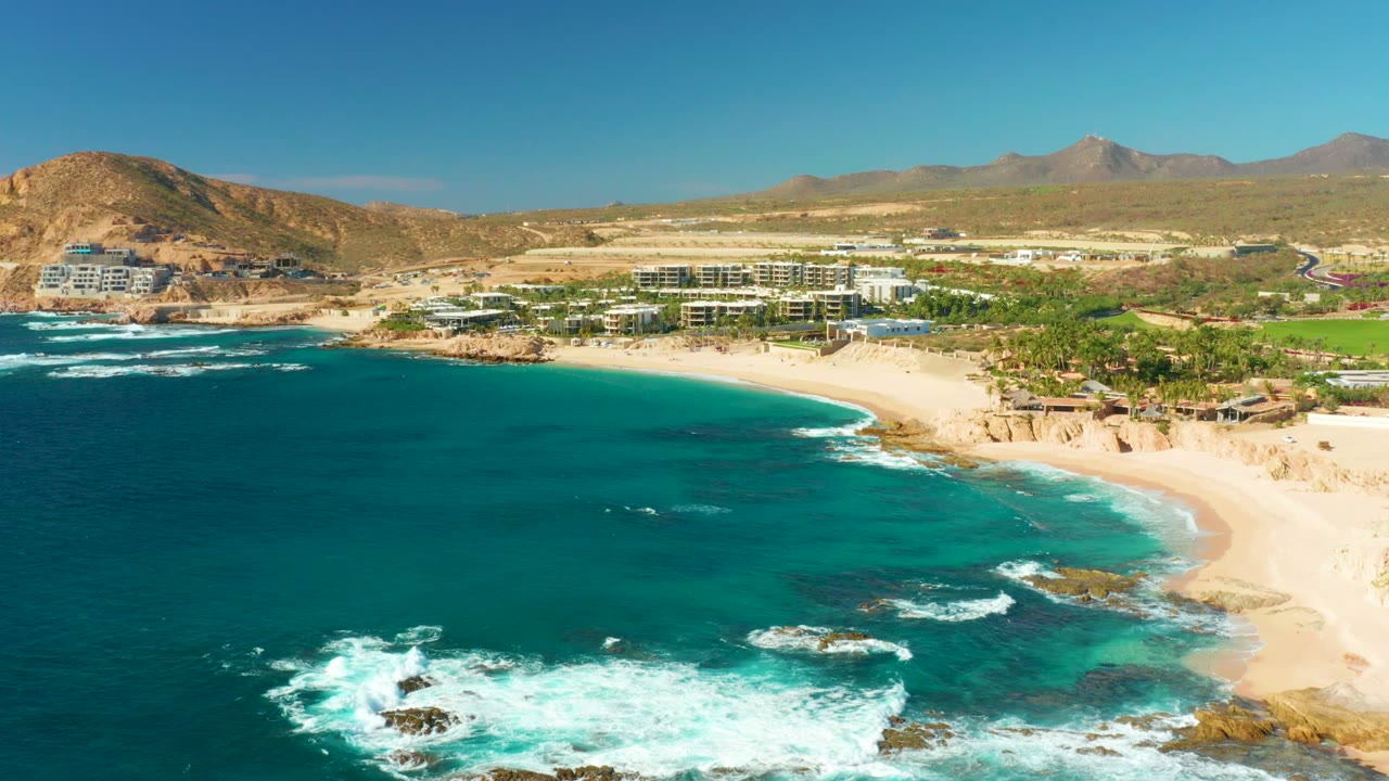 A wide aerial frame of Chileno Beach shows darker blue water at left, foamy surf over rocks, the beach sweeping right, coastal buildings, mountains, and no visible people.