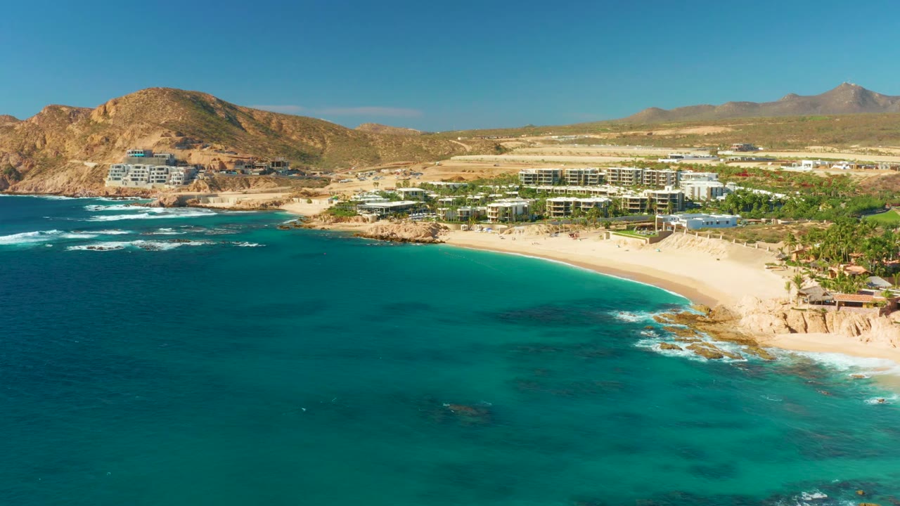 Aerial frame over Chileno Beach in Los Cabos with turquoise water in the foreground, a pale sandy shore, low beachfront buildings, rocky hills, clear sky, and no visible people.
