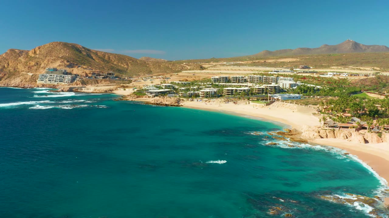 At 0:00, an aerial view of Chileno Beach in Los Cabos shows turquoise water, a curving sandy beach, resort buildings, dry hills, distant mountains, clear sky, and no visible people.