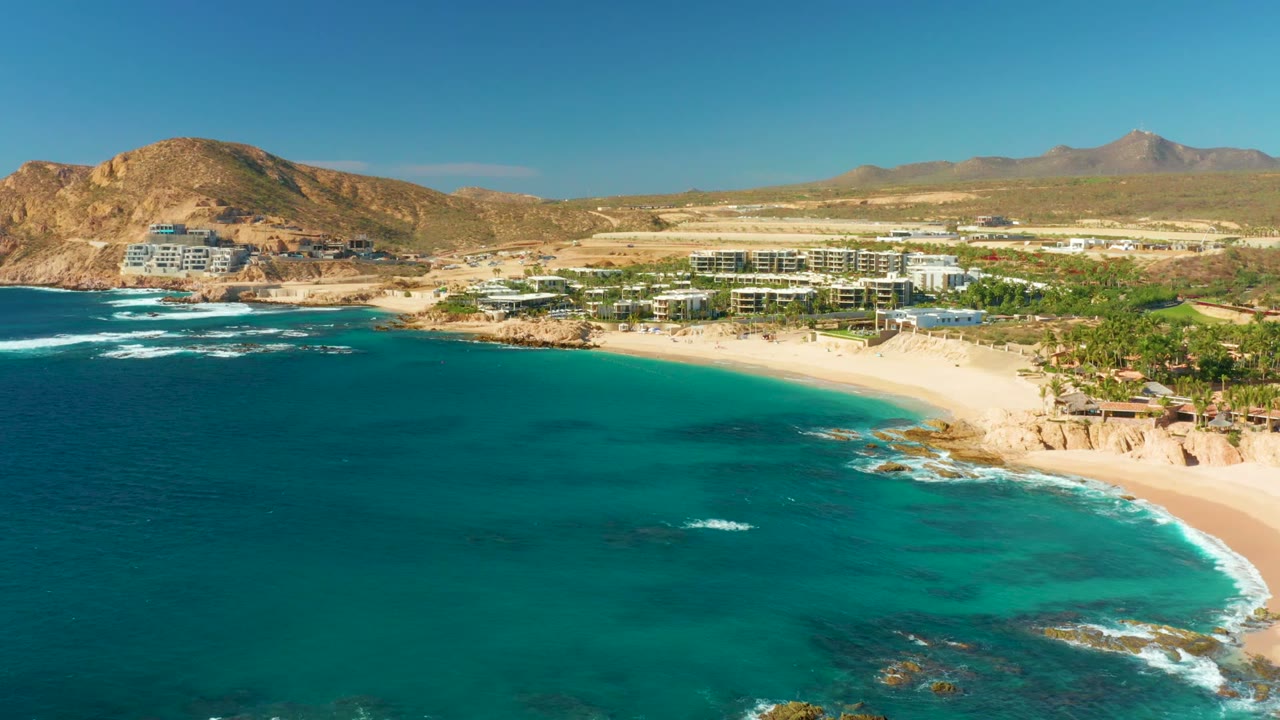Wide aerial coastline with turquoise water, beach, buildings, rocks, hills, and clear sky.