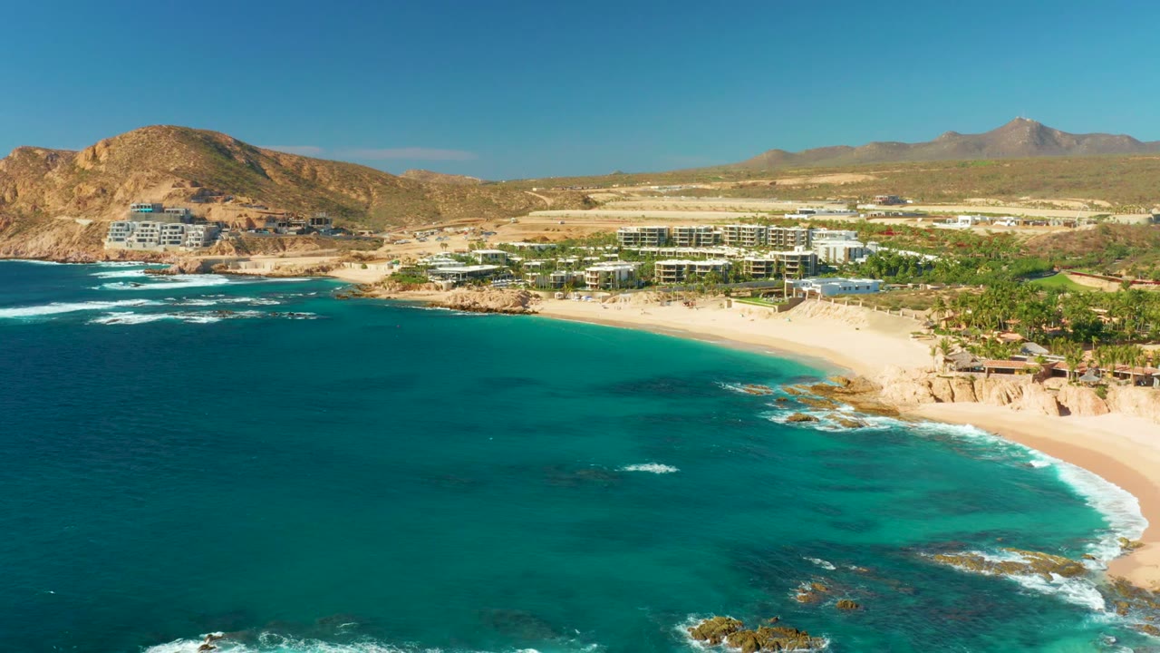 At 0:02, the aerial view shows more open turquoise water in the foreground, wave foam near offshore rocks, the Chileno Beach resort shoreline, desert hills, and no visible people.