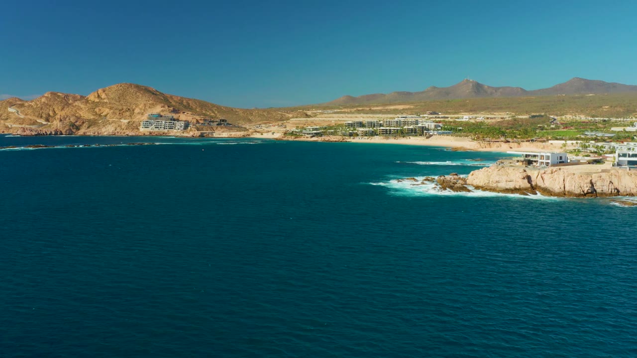 At 0:00, Chileno Beach appears from above with deep blue ocean in the foreground, pale rocks at right, sandy shoreline, low buildings, dry hills, mountains, and no visible people.