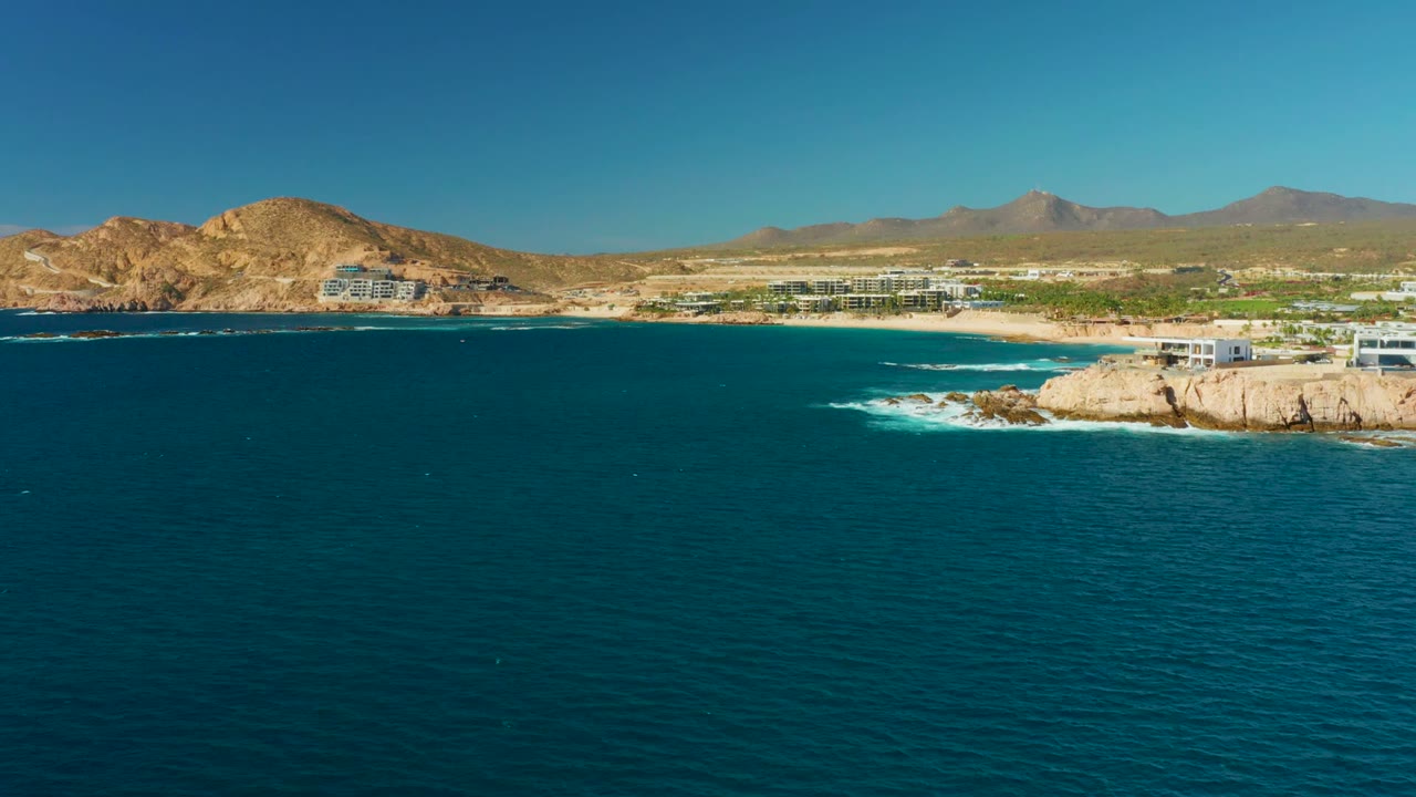 At 0:02, Chileno Beach remains framed wide with dark blue water across the lower frame, foam near the right-side rocks, beachfront buildings, tan hills, mountains, and no visible people.