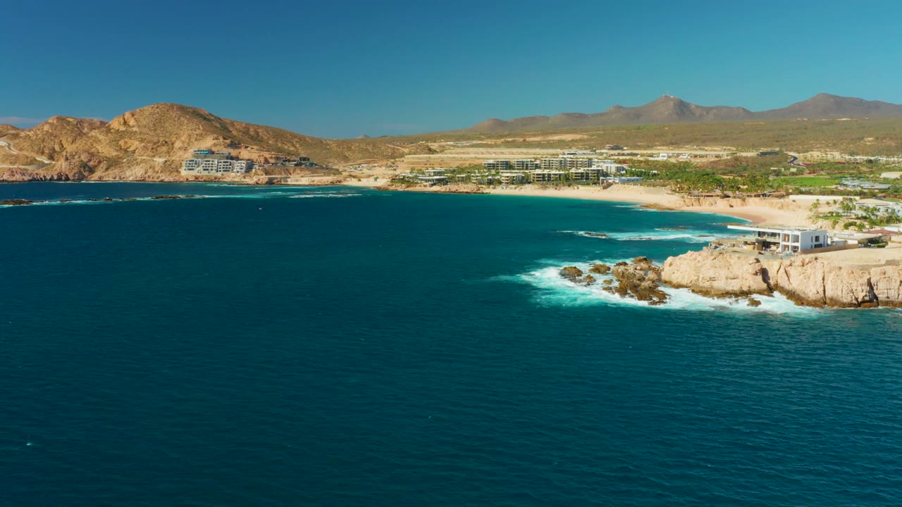Opening aerial frame of Chileno Beach in Los Cabos with deep blue water in front, surf around a rocky point, sandy beach, coastal buildings, dry hills, distant mountains, and no visible people.