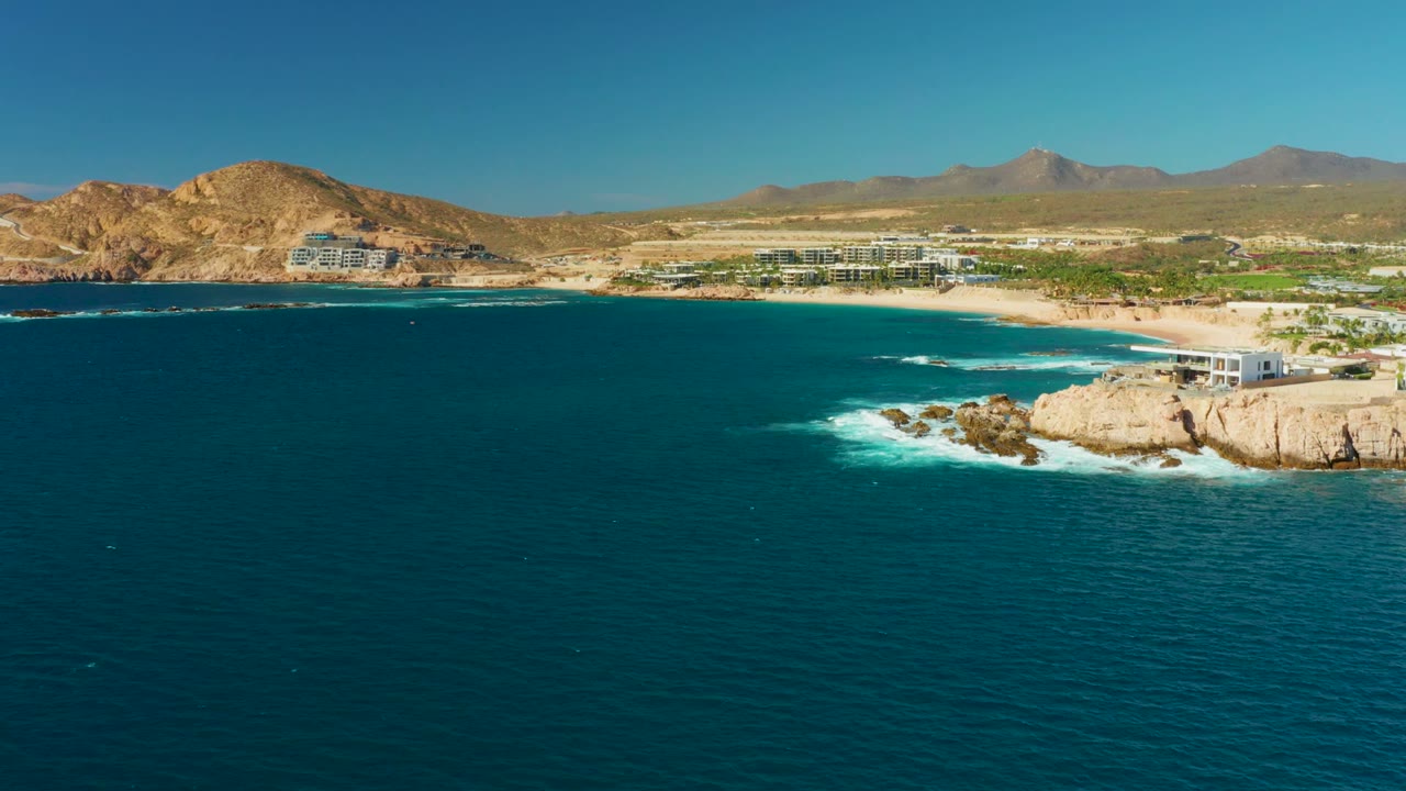 Blue ocean and rocky shoreline with beach buildings below dry hills.