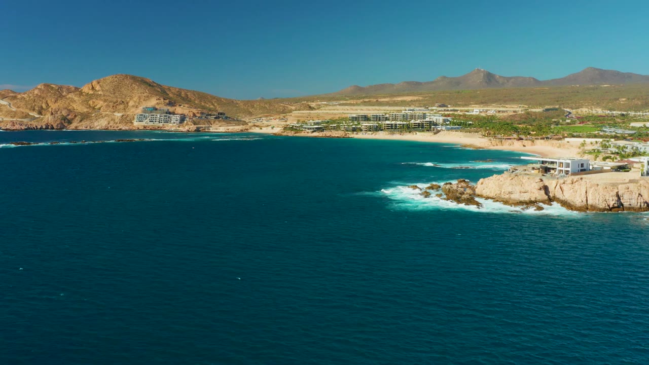 Wide aerial frame of Chileno Beach at 0:02, with open blue water dominating the foreground, a rocky point and white surf to the right, beachfront buildings, tan hills, distant mountains, and no visible people.