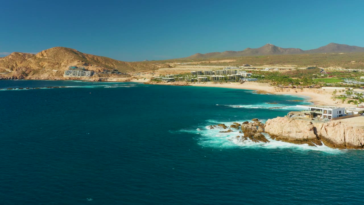 Opening aerial frame over Chileno Beach in Los Cabos, with deep blue water in front, white surf around tan rocks, sandy beach and buildings beyond, distant mountains, and no visible people.