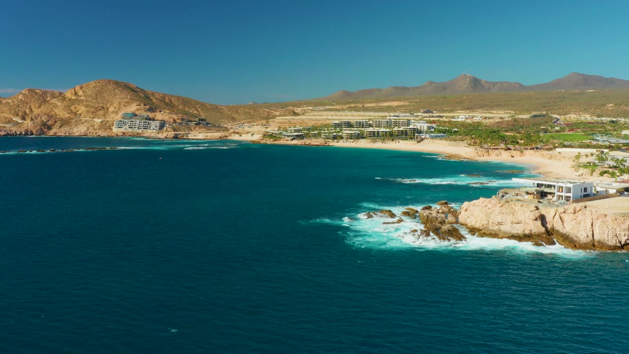 Blue water and white waves near rocks with beachfront buildings and mountains behind.