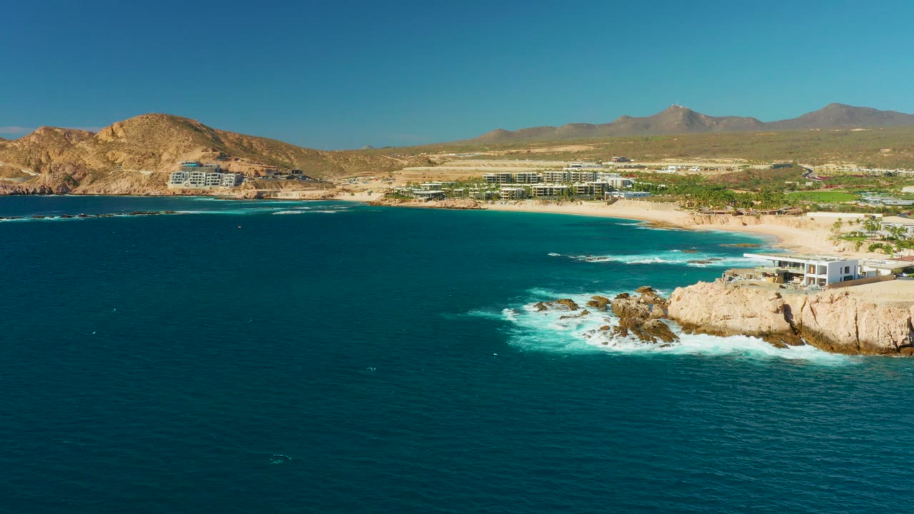 Chileno Beach aerial view with turquoise water close to the shore, white breakers spreading around exposed rocks, beachfront structures on the sand, brown hills behind, and no visible people.