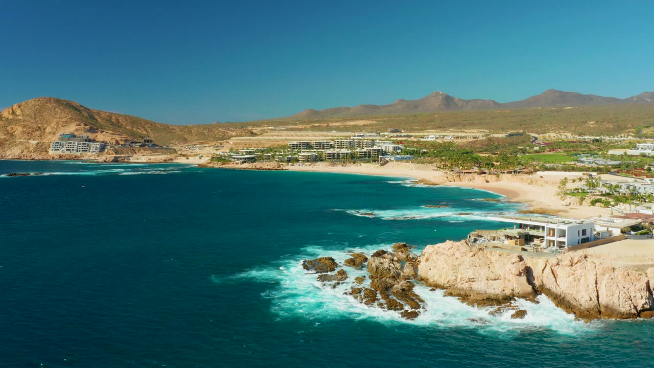 Opening aerial frame over Chileno Beach Los Cabos with deep blue water in front, white surf around rocks at right, sandy beach and low buildings beyond, dry mountains in back, and no visible people.