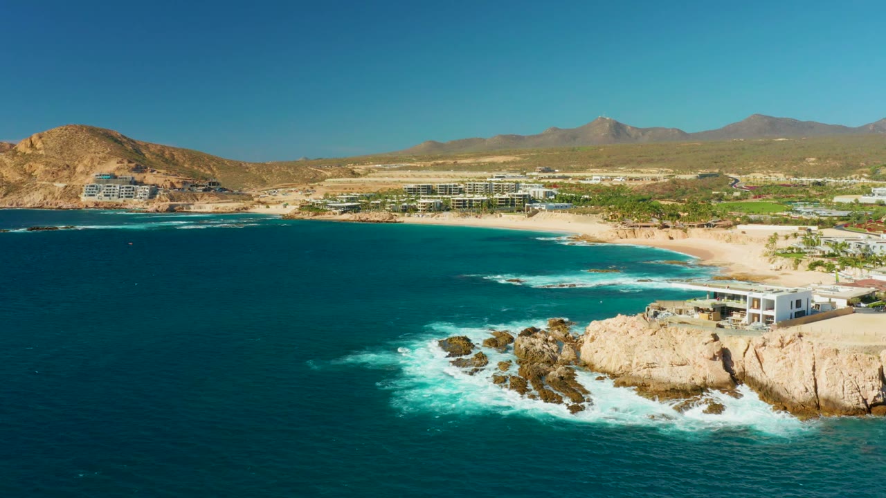 Wide aerial coastline with surf wrapping around rocks and buildings near a sandy beach.