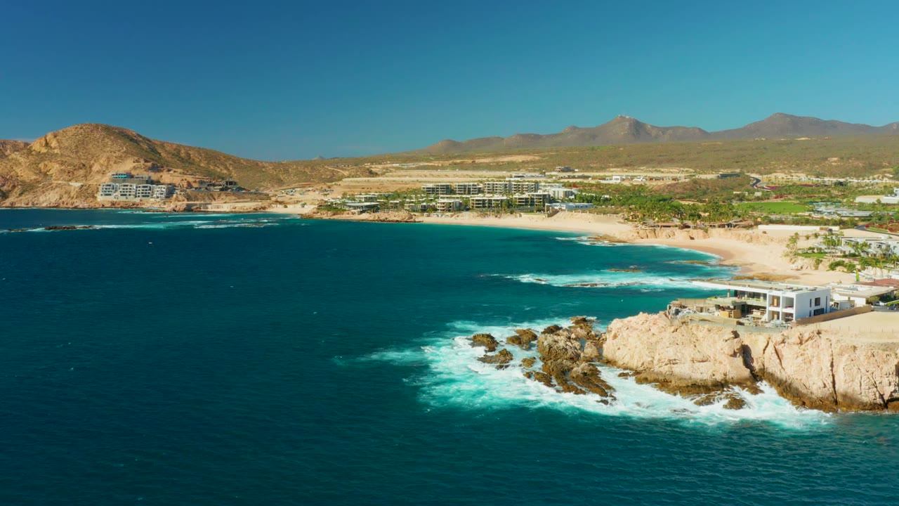 Aerial view of Chileno Beach Los Cabos with darker blue water occupying more of the foreground, white water breaking against tan rocks at lower right, beach development in the distance, mountains, and no visible people.