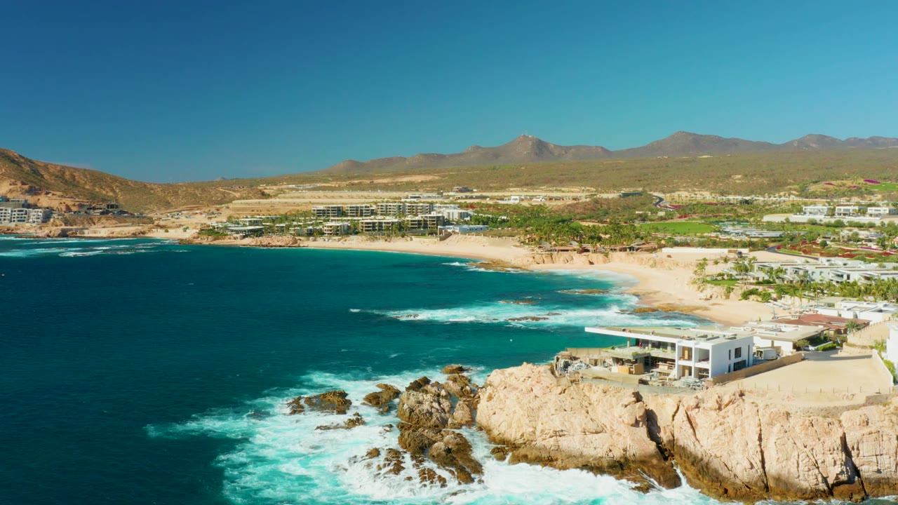 Opening aerial frame of Chileno Beach in Los Cabos with turquoise water, white surf on rocky outcrops, pale cliffs, beachfront buildings, dry hills, distant mountains, and no visible people.
