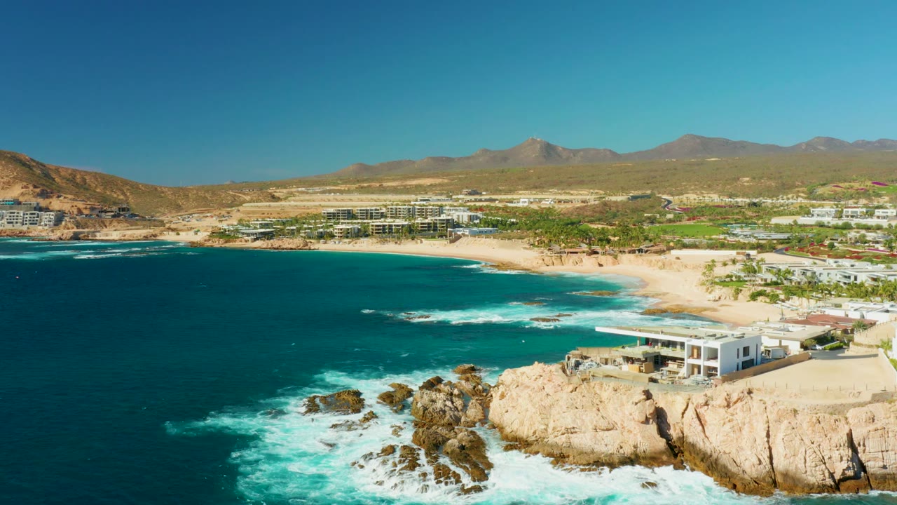 Chileno Beach coastline seen from above, with foamy water spreading around foreground rocks, a broad sand arc and resort-area buildings inland, mountains on the horizon, and no visible people.