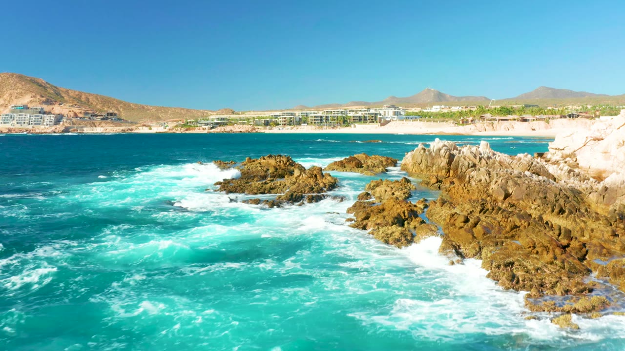 At Chileno Beach in Los Cabos, turquoise water foams around tan shoreline rocks while a sandy beach, low buildings, dry hills, and clear sky sit in the distance, with no visible people.