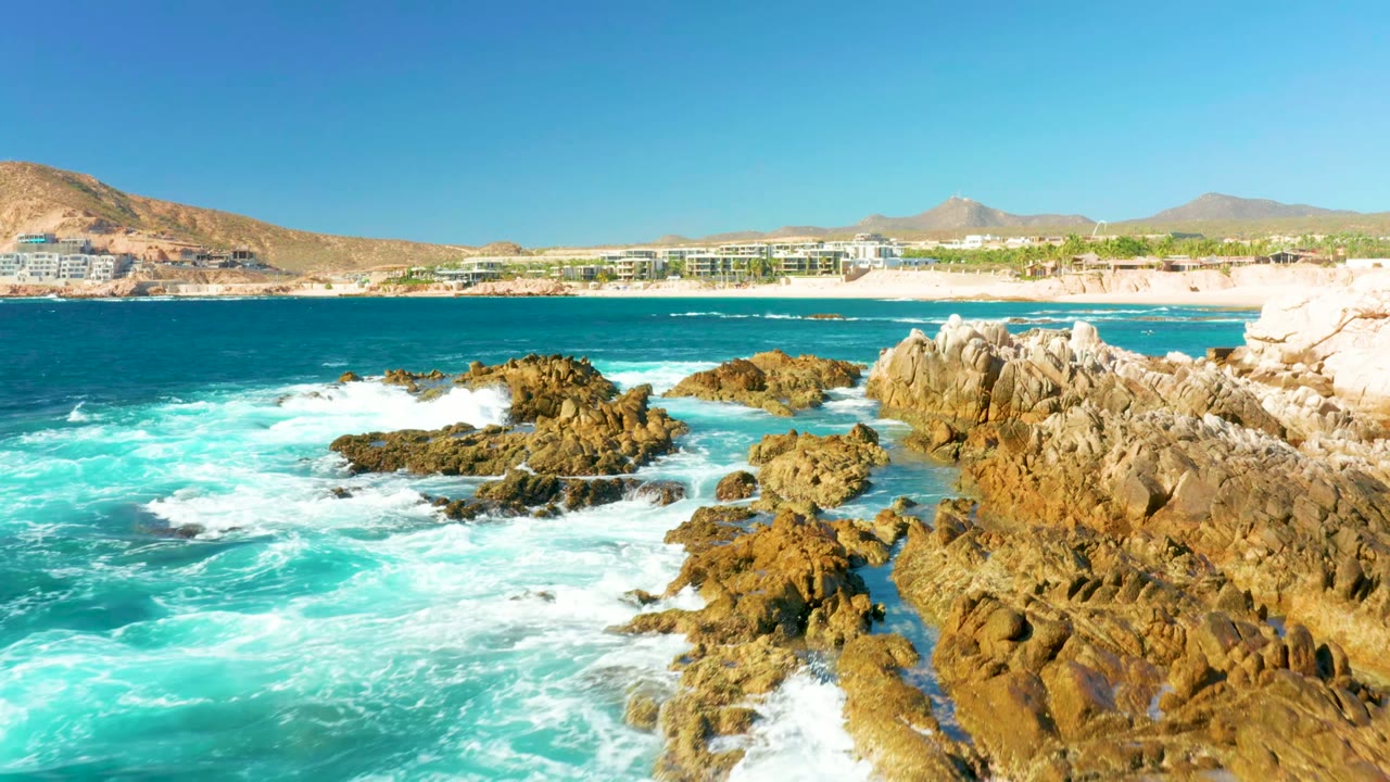 Foamy waves surrounding jagged coastal rocks with beach and hills beyond.
