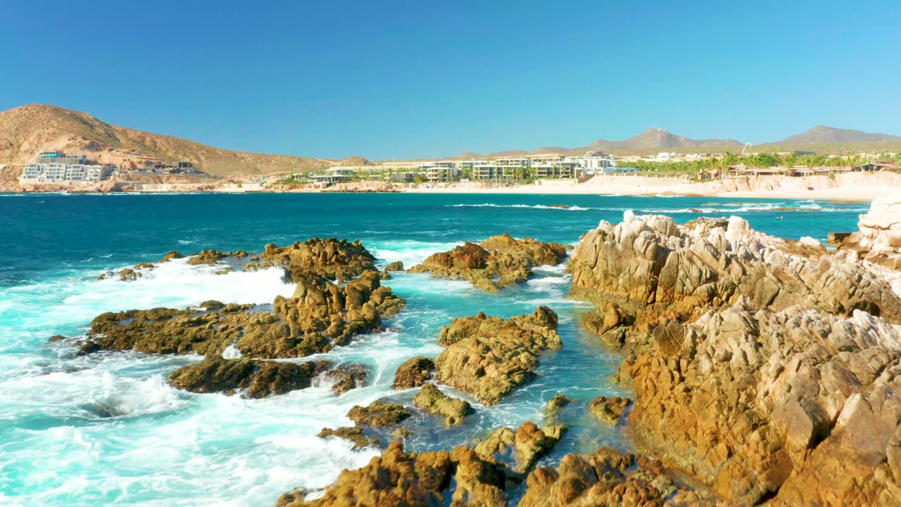 At Chileno Beach in Los Cabos, a rocky outcrop sits near center as surf breaks white around it, with pale beach, low coastal buildings, hills, and a cloudless sky in the background and no visible people.