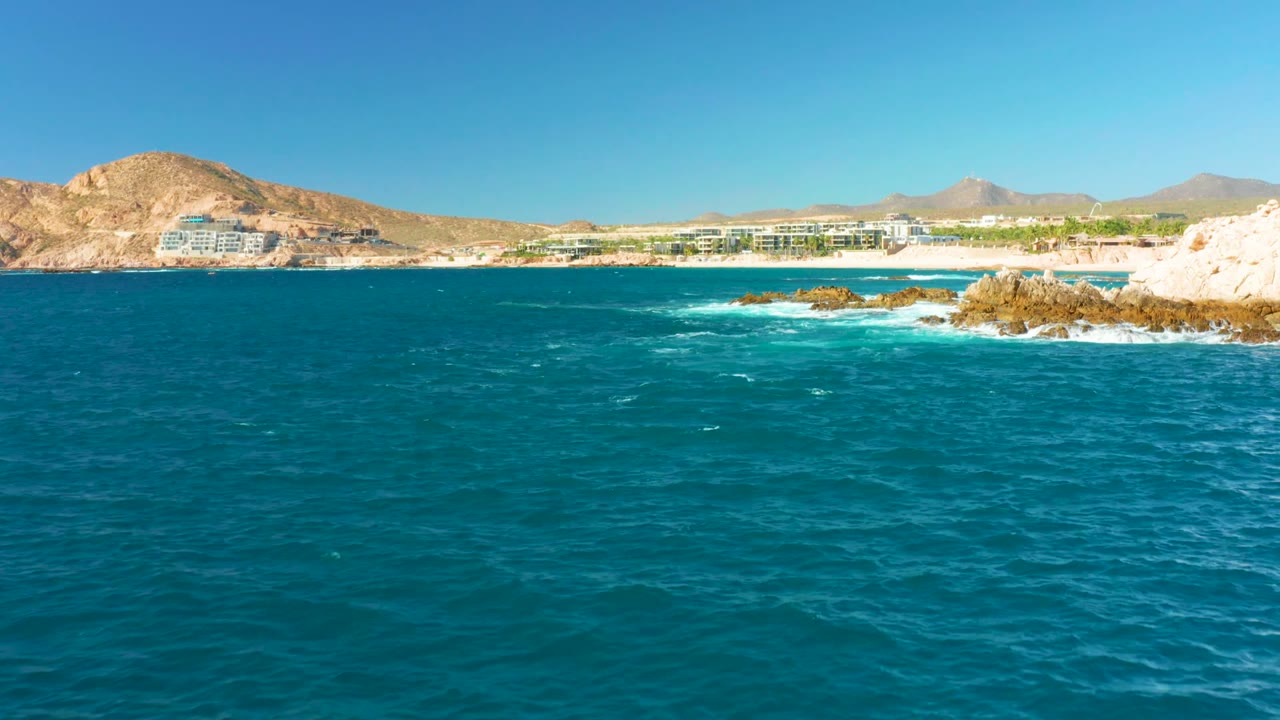Opening frame of Chileno Beach, Los Cabos with turquoise ocean in the foreground, tan rocks at right, distant beachfront buildings and hills, and no visible people.