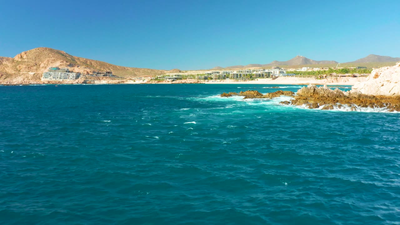 Ocean waves approach low rocks with coastline buildings and hills in the background.