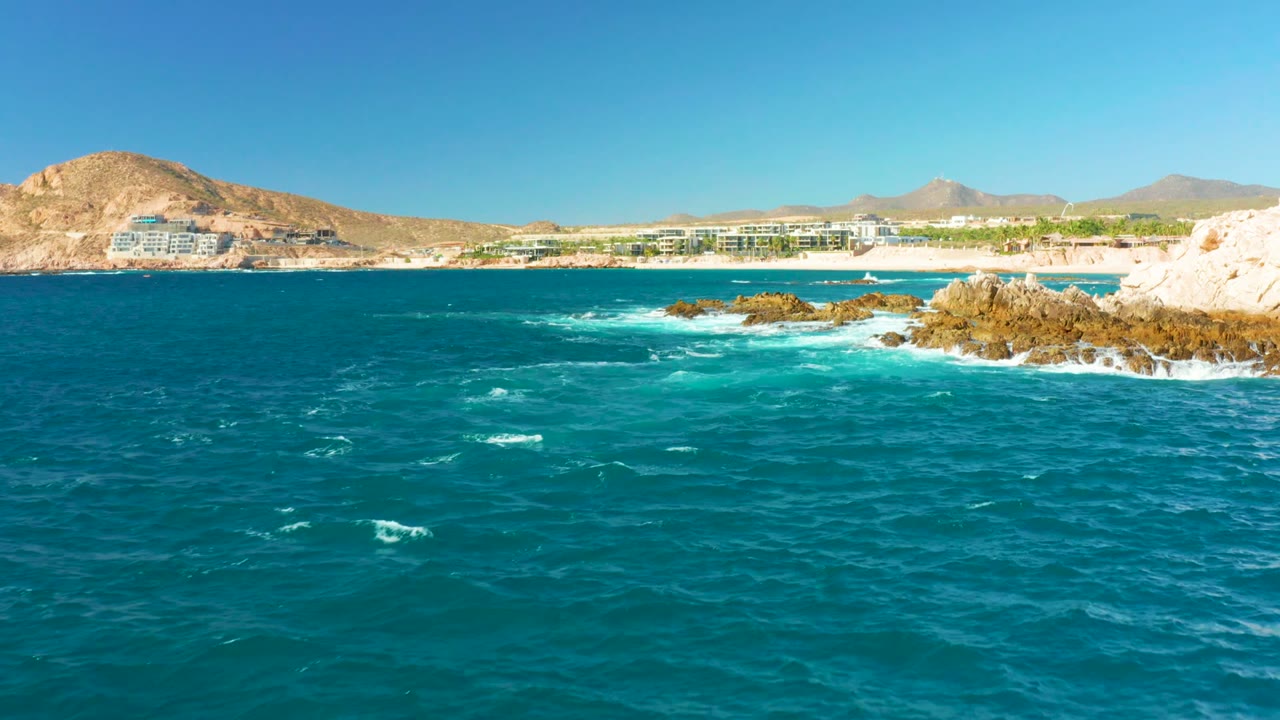 At 0:02, a rocky outcrop on the right frames churning turquoise water, with Chileno Beach buildings and hills across the bay and no visible people.