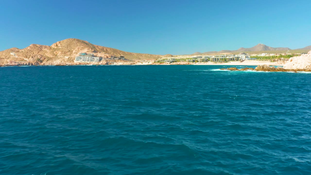Wide view over blue ocean toward Chileno Beach in Los Cabos, with pale rocks, sandy shore, low coastal buildings, tan mountains, clear sky, and no visible people.