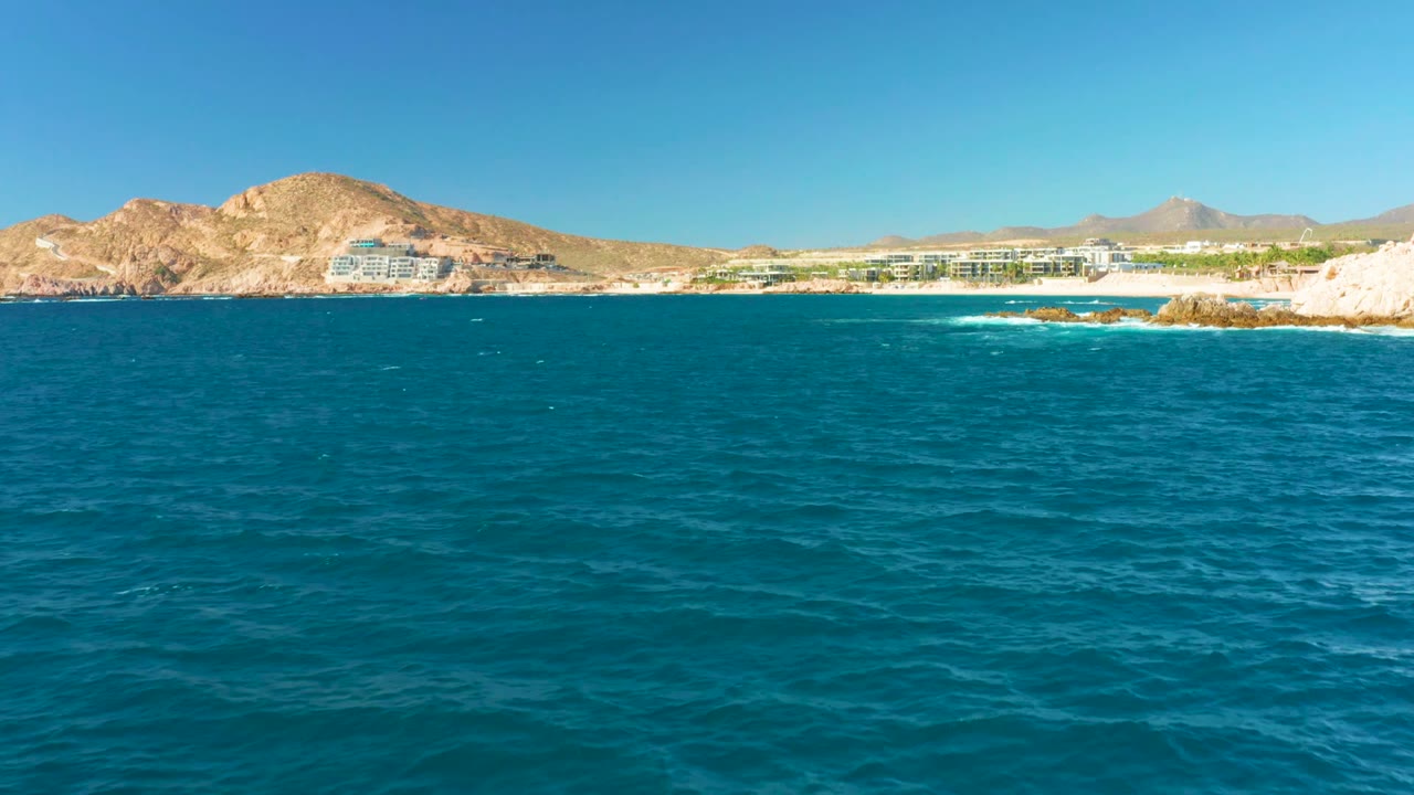 Blue water fills the foreground as small waves move toward pale shoreline rocks at Chileno Beach, with buildings and dry mountains under a clear sky and no visible people.
