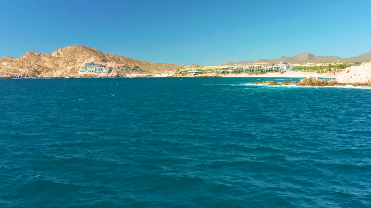 The shoreline sits farther across the blue ocean, with white surf around rocks, beachside buildings below tan hills, distant mountains, clear sky, and no visible people.